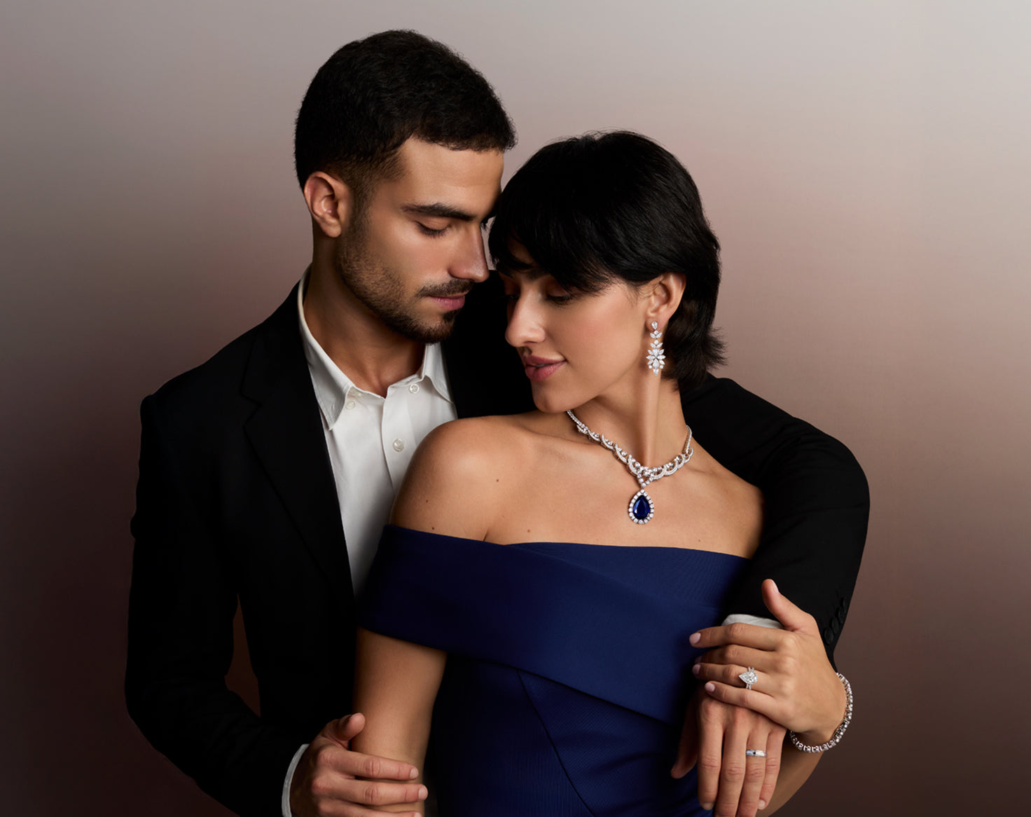 Elegant couple showcasing luxurious diamond and sapphire jewelry. Woman wears a pear-shaped sapphire and diamond necklace, matching diamond drop earrings, a diamond bracelet, and diamond rings, ideal for engagements, weddings, and anniversaries.