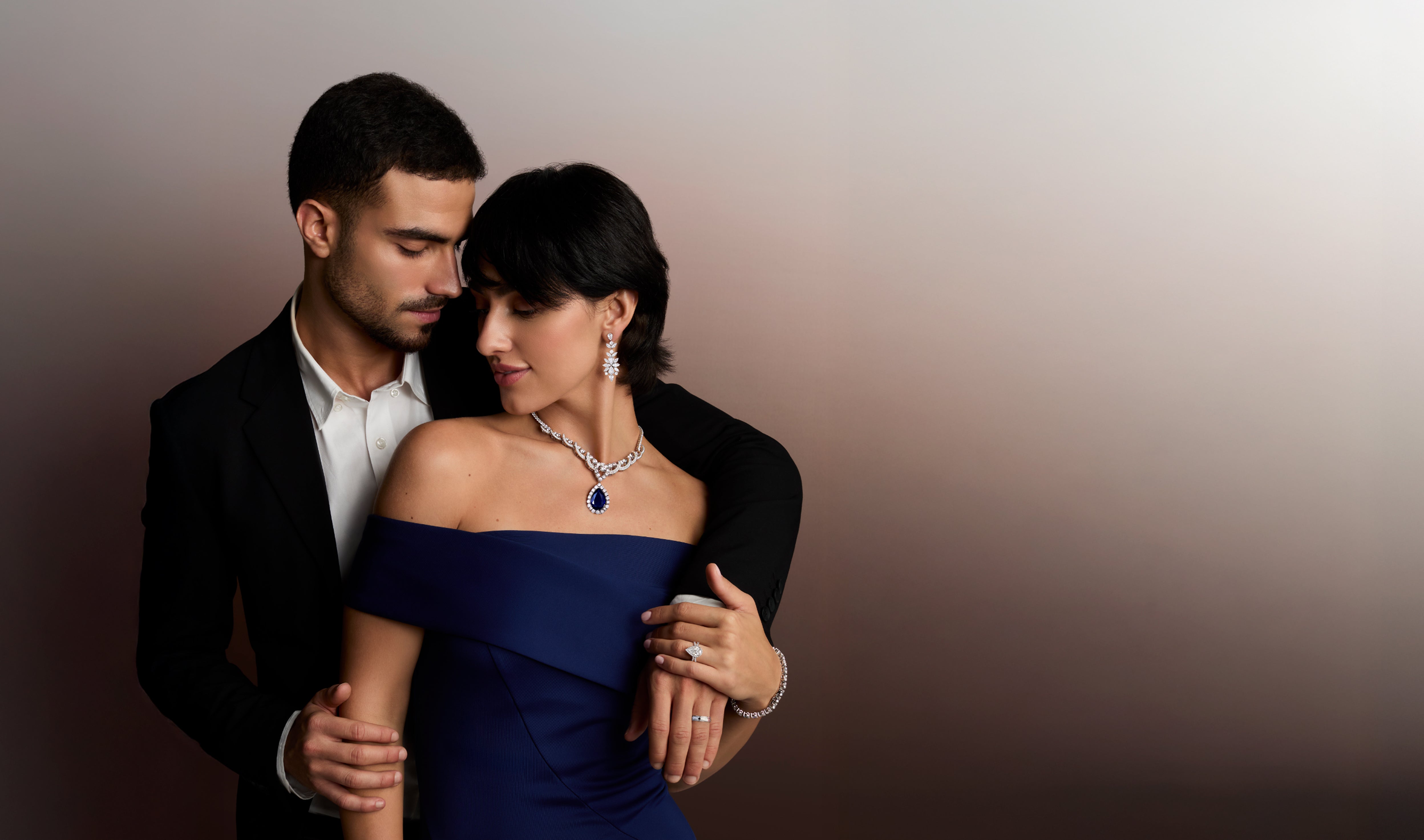 Elegant couple showcasing luxury diamond jewelry: a woman wears a dazzling diamond and sapphire necklace, diamond drop earrings, a diamond tennis bracelet, and diamond rings, embraced by a man in a suit. Ideal for engagement rings, wedding bands, and fine diamond collections.