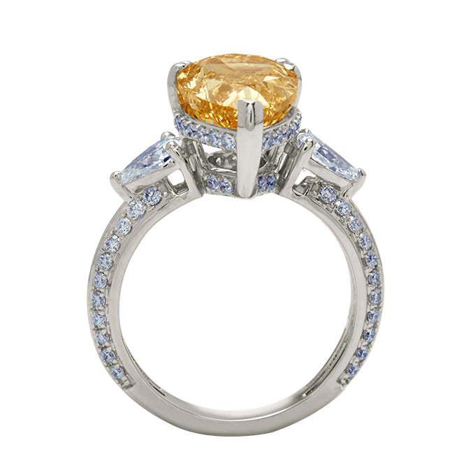 The Champagne Flame Pear Ring - a luxurious diamond engagement ring featuring a radiant champagne pear-cut diamond, flanked by white pear diamonds, with a delicate blue diamond halo and a pavé diamond white gold band.
