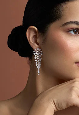 A close-up of a woman wearing a dangling teardrop-shaped silver earring adorned with clear stones, set against a warm terracotta background.