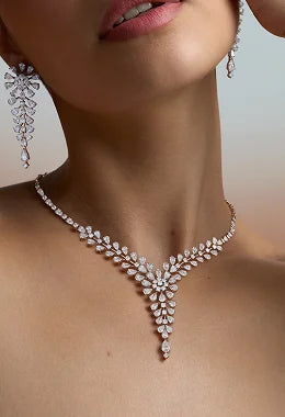 A close-up of a model wearing an ornate silver-toned Y-shaped necklace with matching drop earrings. The jewelry features intricate floral and leaf-like designs set with sparkling clear stones, shown against the model's neck and upper chest.