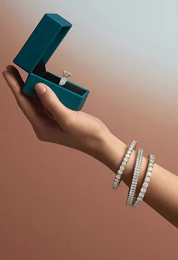 Hand holding a teal jewelry box with an engagement ring, wearing silver bracelets on a gradient background
