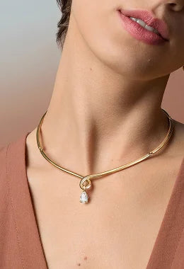 A close-up of a model wearing a gold chain necklace with a small teardrop-shaped clear gemstone pendant, styled with a terracotta top against a soft beige background.