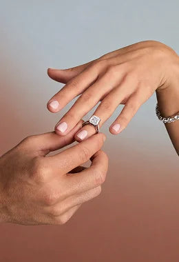 A close-up of hands wearing an engagement ring. The ring features a square-cut clear gemstone set in a four-prong silver or white gold setting.